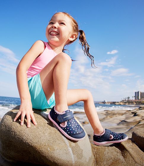 A girl at the beach sitting on a boulder smiling.